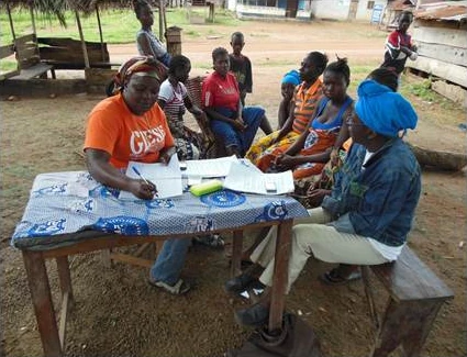 A community health worker registers households in a village register while women gather around the table, rural Liberia