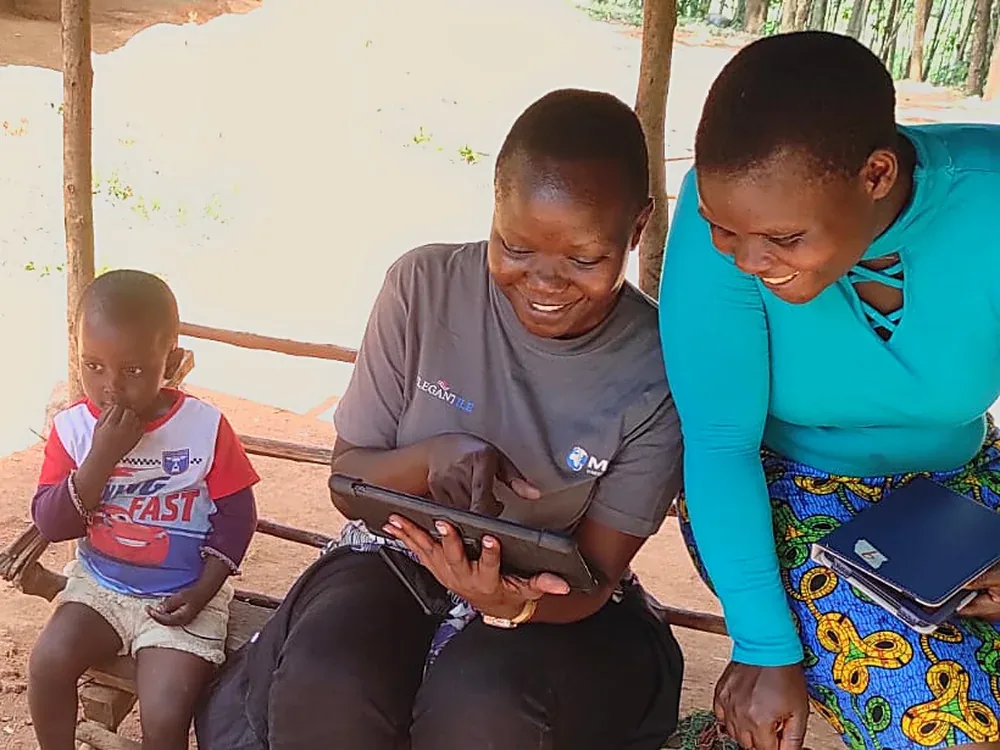 A community health worker records a household visit on a tablet while a mother looks on, rural Kenya